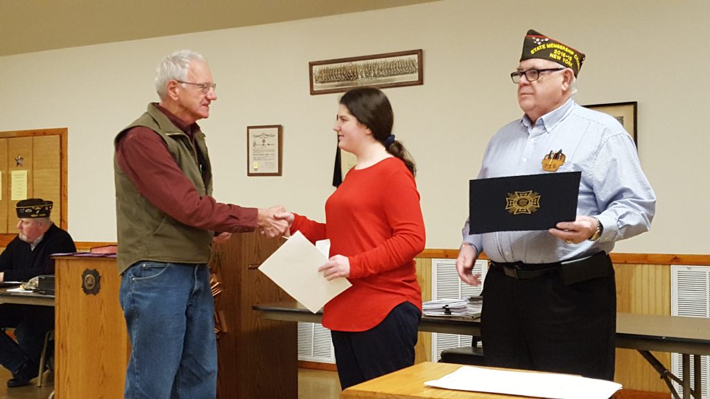 middle school student shakes hands with a representative of the VFW while a second representative, at right, holds a certificcate