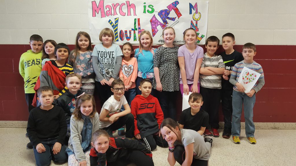 third grade class stands before a banner that says "march is youth art month" in a school hallway