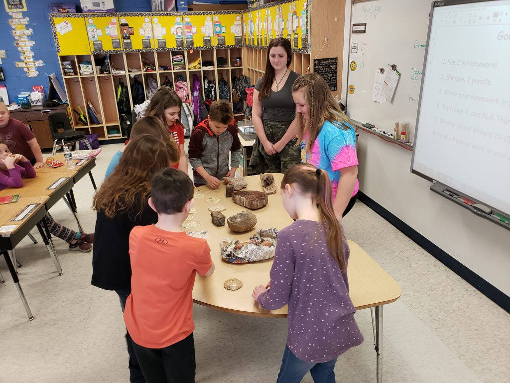 elementary students observe fossil collection on table with high school students