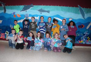 A dozen elementary students stand in front of a mural depicting an ocean scene