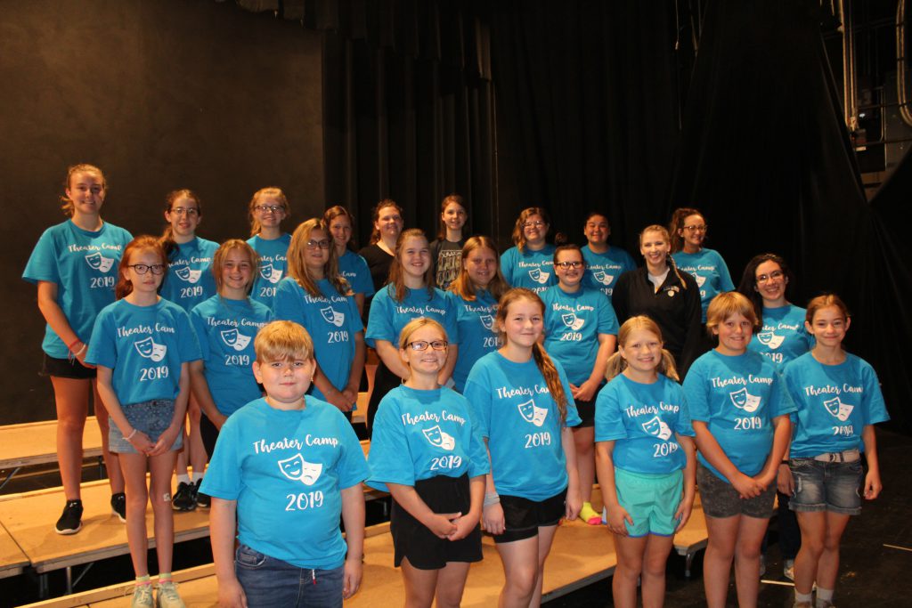 Group of students wearing blue theater camp t-shirts stand on risers on a stage in a school auditorium