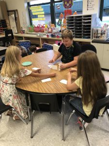 three students seated at table in elementary classroom build a card tower