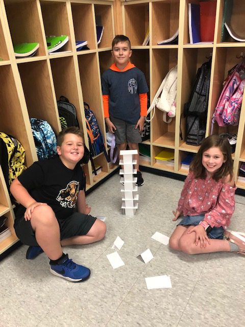 three students stand before cubbies behind a card tower they made in an elementary classroom