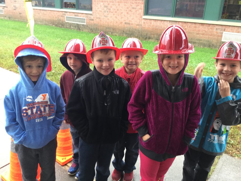 group of six students wearing plastic firemen hats outside of an elementary school