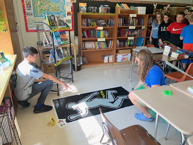 students kneel next to a mock crime scene in a middle school classroom