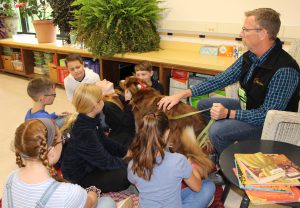 teacher sits in a wicker chair before students seated on a carpet with a therapy dog in a classroom