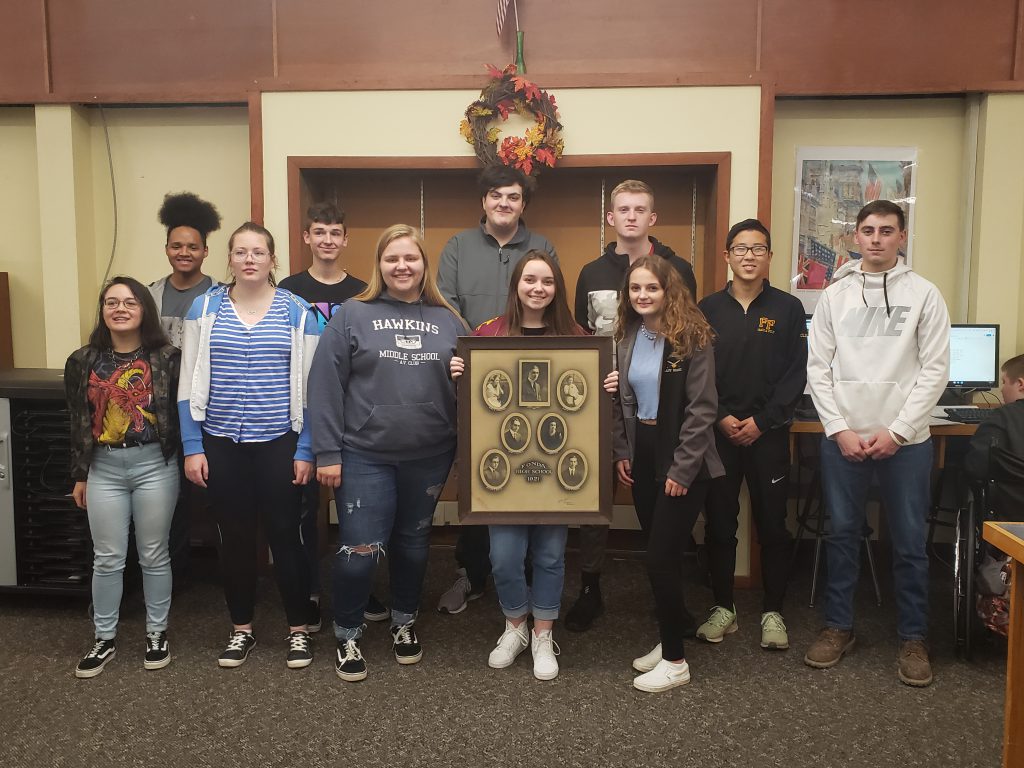 group of high school students pose in a library while holding a picture of the class of 1921