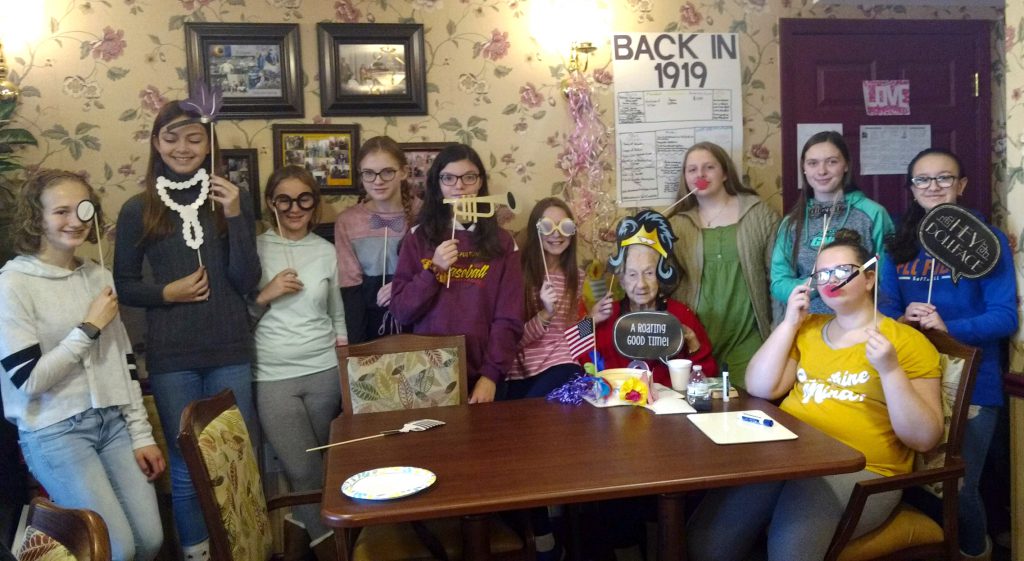 middle school students and a nursing home resident seated around a table holding photo booth props