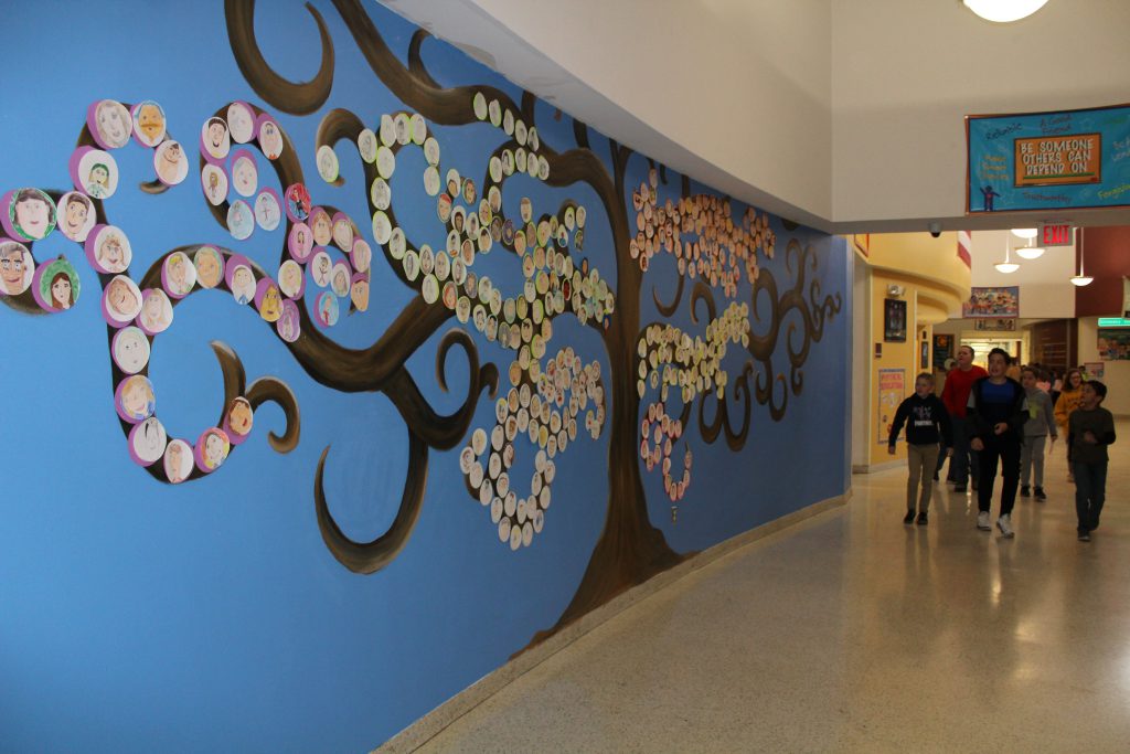 students walk by a painting of a large tree on a wall in an elementary school foyer