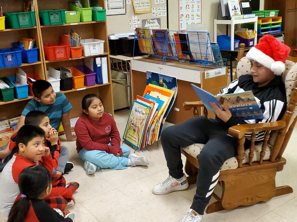 high school student seated in a rocking chair reads to elementary students seated on a classroom floor
