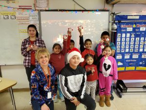 group of elementary students, their teacher and a high school student wearing a santa hat pose for a picture in front of a classroom