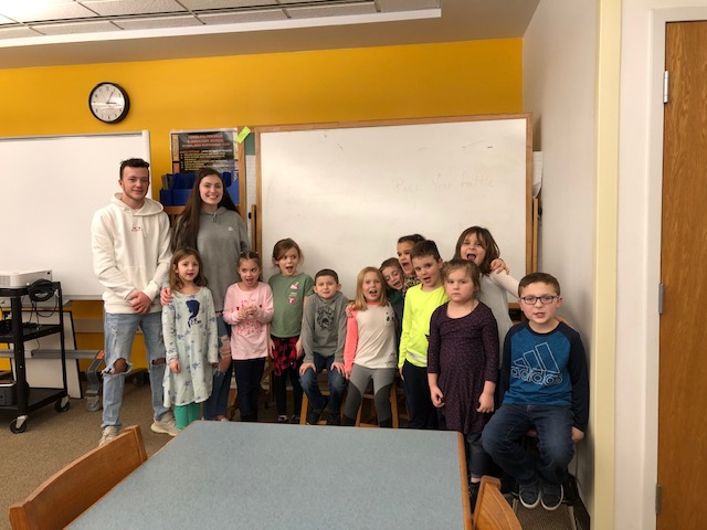 two high school students stand with a group of children in a school classroom