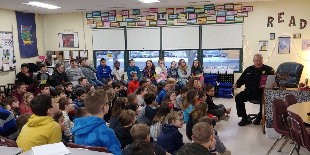 law enforcement official in uniform at the front of a classroom reading to a large group of middle school students