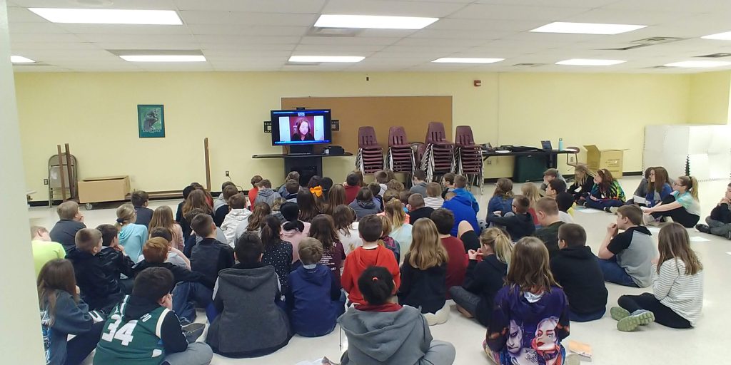 group of students seated on a floor in front of a screen displaying a video chat