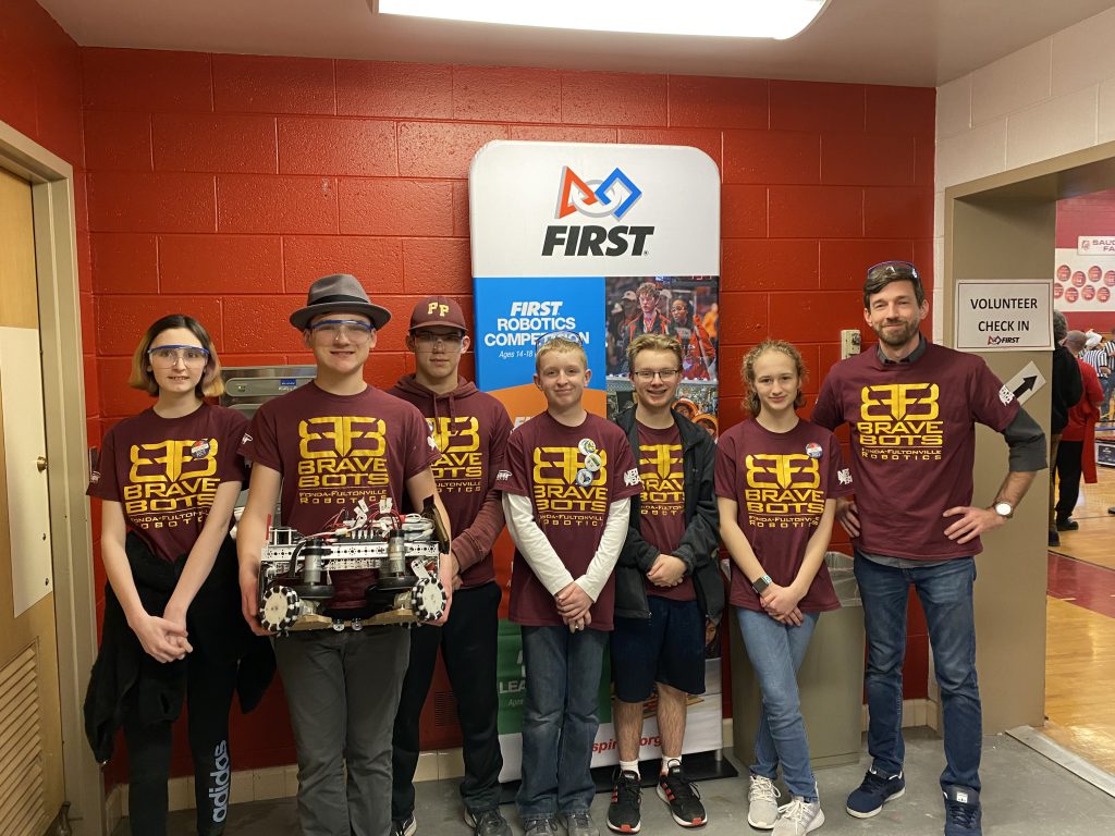 six students and a coach wearing Brave Bots T-shirts in front of a FIRST robotics banner