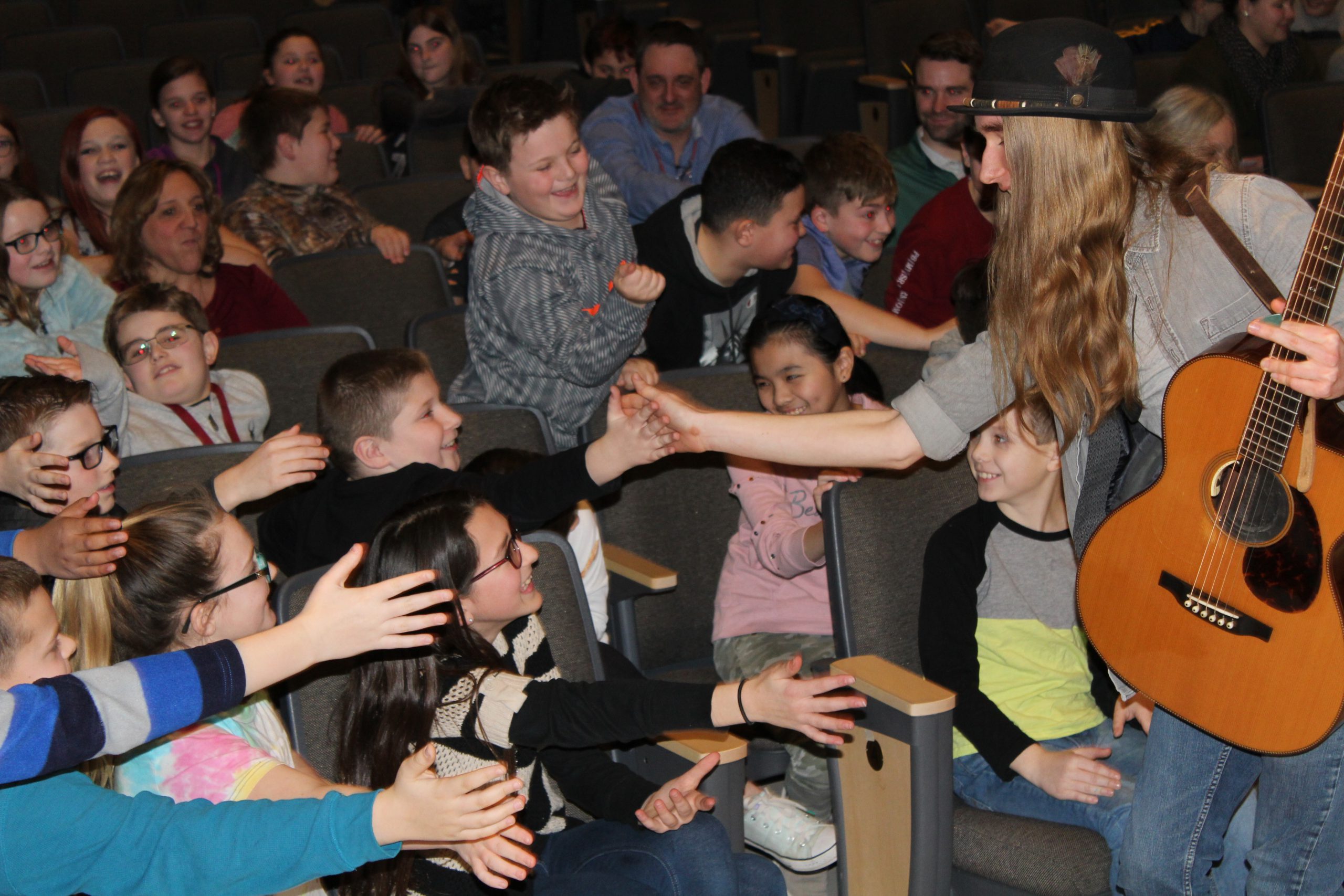 musician holding a guitar gives middle school students high fives in a school auditorium