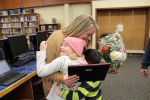 two children hug a person holding a plaque and flowers