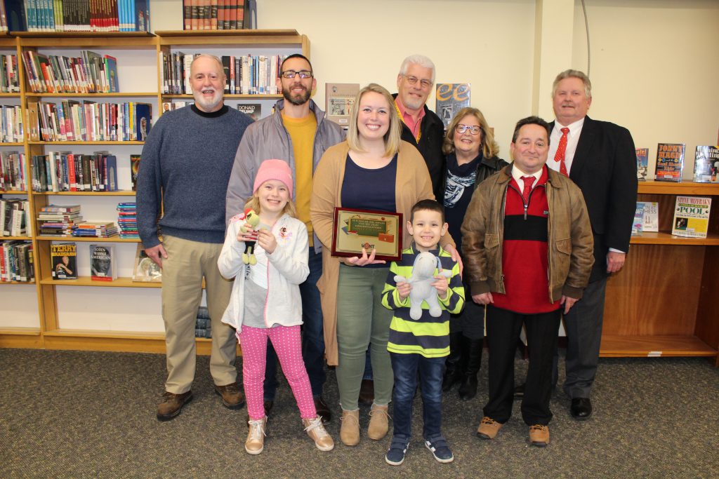 a teacher holds a plaque while flanked by family members and radio station employees in a school library