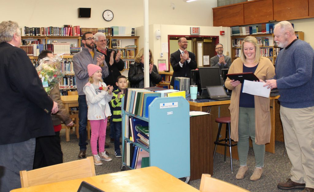 teacher is presented with a plaque from a radio dj in a school library while surrounded by school staff and family members