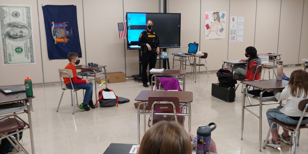 adult in a police uniform stands at the front of a middle school classroom as students are seated in desks