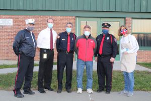 three firefighters, a school superintendent, a community volunteer and a teacher all wearing face masks posing for a photo outside of a school building.