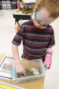 second grade student wearing a mask points inside a diorama built in a plastic tote
