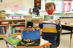 student wearing a mask holds up a paper while standing next to a diorama of a snake habitat