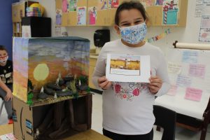 student holds up a paper while standing next to a diorama in a classroom