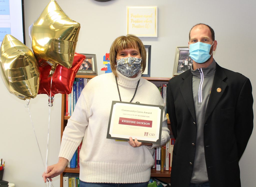 a school pupil services director with shoulder length hair wearing a mask holds a bunch of balloons and an award certificate while standing next to a school superintendent in an office