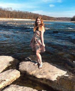 high school student with long hair wearing a dress stands on a rock in a body of water 