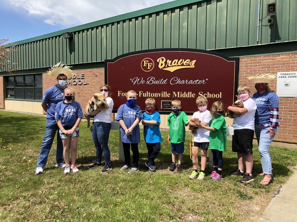 a group of elementary and middle school students stand with their principals outside of the school while holding baby pigs